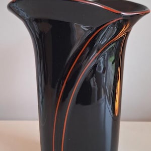 Op de afbeelding: Een zwarte vaas met een uitlopende bovenkant en oranje accentlijnen. De vaas heeft een glanzende afwerking en een modern design. Het is een decoratief item voor in huis.