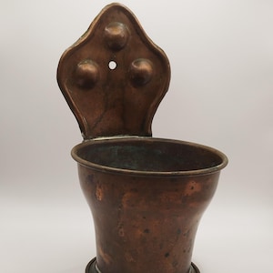 May include: An antique copper wall-mounted container, featuring a flared body and a decorative backplate. The backplate has three raised circular details and a small hole. The container shows a dark patina, indicating age and use.