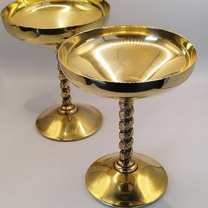Op de afbeelding: Twee goudkleurige champagnecoupeglazen. Elk glas heeft een brede, ondiepe kom en een getextureerde steel. De basis van elk glas is rond. De glazen worden op een wit oppervlak gepresenteerd.