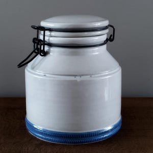 Pode incluir: Um pote de cerâmica branco com tampa e fecho de metal preto. A base do pote tem uma faixa decorativa azul. O pote é cilíndrico com uma base ligeiramente mais larga e um pescoço mais estreito.