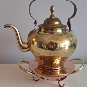 Peut inclure: Une théière vintage en laiton avec un chauffe-plat en cuivre. La théière a un corps arrondi, un bec courbé et une poignée décorative. Le chauffe-plat a une base circulaire avec des poignées incurvées et de petits pieds.