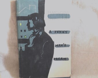 May include: A hardcover book titled A Funny Man's Dream by Dostoevsky. The cover features a black silhouette of a person standing against a blue building background with yellow windows. The text is printed in black on a white background.