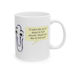 May include: White ceramic mug with a cartoon paperclip character and googly eyes. The mug has a speech bubble with the text: "It looks like you're about to fuck around. Would you like to find out?"
