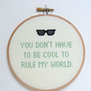 Puede incluir: Un aro de bordado blanco con un diseño de punto de cruz verde que dice "You don't have to be cool to rule my world." El diseño también incluye un par de gafas de sol negras.