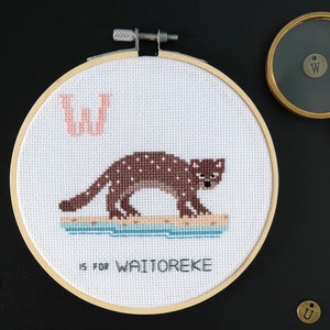May include: A white embroidery hoop with a brown and white illustration of a weasel on a blue and white background. The letter "W" is stitched in pink above the weasel. The text "W is for Waitoreke" is stitched below the weasel.
