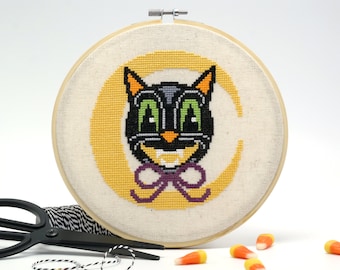 May include: A black cat with green eyes and a purple bow tie is stitched on a white fabric background. The cat is in a yellow crescent moon shape. The fabric is stretched over a wooden embroidery hoop.