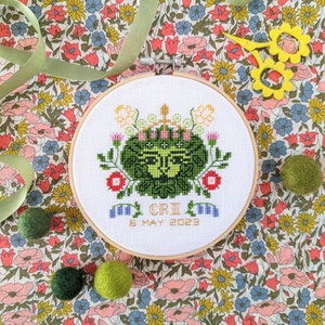 May include: A white embroidery hoop with a green, yellow, and red floral design. The design features a green face with a crown of flowers. The text "C.R.I. 5 MAY 2023" is embroidered below the design.