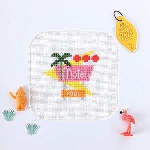 May include: Cross-stitch pattern of a pink motel sign with a palm tree and the words "motel" and "pool". The sign is yellow and white with red circles. The pattern is on a white background.