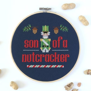 May include: A blue embroidery hoop with a cross-stitch design of a nutcracker soldier with the text "son of a nutcracker" in red and green thread.