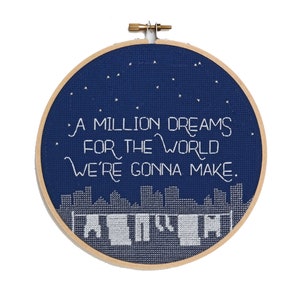 May include: A circular navy blue cross-stitch embroidery hoop featuring the text "A Million Dreams For The World We're Gonna Make." The design includes a cityscape and stars, all in white thread. The hoop is made of wood.