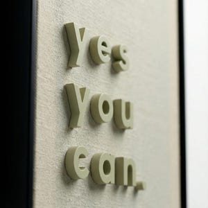 May include: Close-up of a framed artwork with the words "Yes you can." in 3D, light green letters. The letters are mounted on a textured, off-white canvas background, framed by a black border.