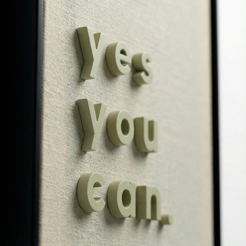 May include: Close-up of a framed artwork with the words "Yes you can." in 3D, light green letters. The letters are mounted on a textured, off-white canvas background, framed by a black border.