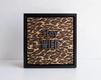 May include: Square framed artwork with a leopard print background. The words "stay WILD" are in black, raised lettering. The frame is black, and the artwork is displayed on a white surface.
