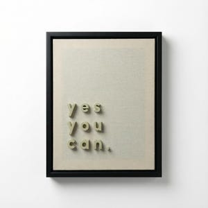 May include: Framed artwork with the words "yes you can" in 3D lettering. The text is in a light green color and is placed on a beige canvas. The frame is black and the background is white.