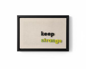 May include: A framed canvas with the words "keep strange" in black, green, and yellow letters. The canvas has a neutral beige tone and is set within a black frame. The artwork is a modern, minimalist design.
