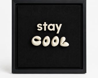 May include: A square black frame containing a black fabric background with the words stay COOL in raised, off-white, rounded, three-dimensional lettering. The word stay is positioned above the word COOL, which features a stylized, bubbly font.