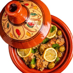 Pode incluir: Um tajine marroquino tradicional, um pote de barro com tampa cónica, está parcialmente aberto, revelando um prato de frango e azeitonas guarnecido com rodelas de limão e coentros. O tajine é castanho-avermelhado com padrões decorativos na tampa.
