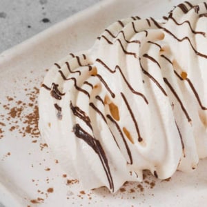 May include: A close-up of a dessert on a white rectangular plate. The dessert is white with dark brown chocolate drizzle. Cocoa powder is sprinkled around the dessert on the plate. The dessert has a layered, textured appearance.