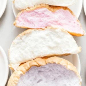 Puede incluir: Primer plano de merengues coloridos cortados en rodajas. Los merengues, presentados en platos blancos, revelan capas de rellenos rosa, blanco y morado. Las cáscaras de merengue son de color marrón dorado, creando un contraste llamativo.