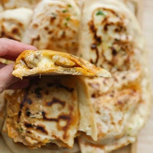 May include: A close-up of a hand holding a piece of freshly baked flatbread, revealing a savory filling. The bread has a golden-brown crust with dark spots, and the filling appears to be a mix of cheese and other ingredients. The food is on a wooden surface.