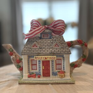 May include: A ceramic teapot shaped like a cottage with a red door, blue siding, and a grey shingled roof. A sign reads APPLE COTTAGE. The handle and spout resemble tree branches with red apples. A red and white striped ribbon is tied on top.