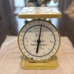 May include: A vintage yellow Sears kitchen scale with a round dial face. The dial displays measurements in pounds up to 25 pounds and kilograms up to 11 kilograms. The text reads Sears, Weighs 25 Pounds by Ounces, 11 kilograms by 50 grams, NOT LEGAL FOR TRADE.