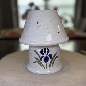 May include: A ceramic candle warmer with a speckled white glaze and a flared top section featuring small ventilation holes. The cylindrical base is decorated with a hand-painted blue iris flower and green leaves on a speckled white background.