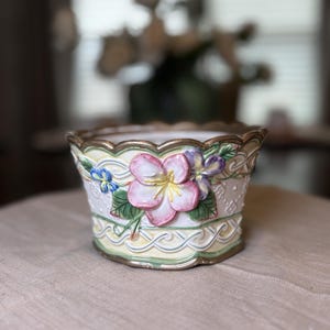 Peut inclure: Un cache-pot en céramique à bord festonné, présentant une base de couleur crème avec des détails floraux en relief, notamment des fleurs roses et violettes, des feuilles vertes et un motif en treillis jaune clair sur le bord inférieur doré.