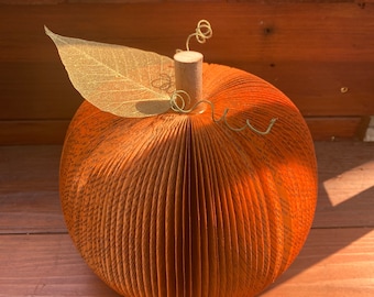 Puede incluir: Una decoración de calabaza de papel naranja hecha a mano. La calabaza está hecha de páginas de libro dobladas, con un tallo de madera, una hoja dorada y una vid de alambre. La calabaza está sobre una superficie de madera.
