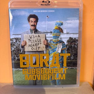 May include: A Blu-ray disc case for the movie "Borat Subsequent Moviefilm" featuring Sacha Baron Cohen. The cover shows Cohen holding a sign that reads "US+A PLEASE ACCEPT GIFT". The title is in large, gold letters.