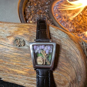 May include: A rectangular wristwatch with a black leather strap. The silver watch face features a miniature scene with purple flowers, a small log, and a tan object. The watch is displayed on a piece of wood.