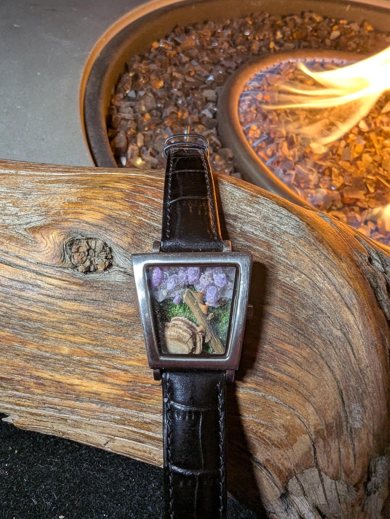 May include: A rectangular wristwatch with a black leather strap. The silver watch face features a miniature scene with purple flowers, a small log, and a tan object. The watch is displayed on a piece of wood.