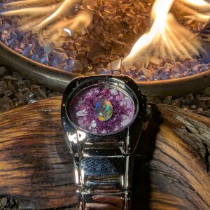 May include: A silver wristwatch with a round face filled with purple and clear stones and a central iridescent orb. The watch is set against a backdrop of flames and glass rocks, with a wooden surface.