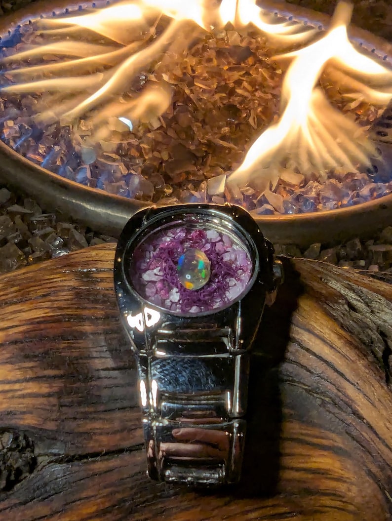 May include: A silver wristwatch with a round face filled with purple and clear stones and a central iridescent orb. The watch is set against a backdrop of flames and glass rocks, with a wooden surface.