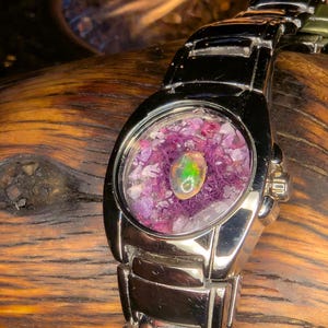 May include: A silver wristwatch with a unique face. The watch face is clear, revealing a colorful arrangement of small purple stones and a central opal. The opal has green and yellow hues. The watch band is also silver.