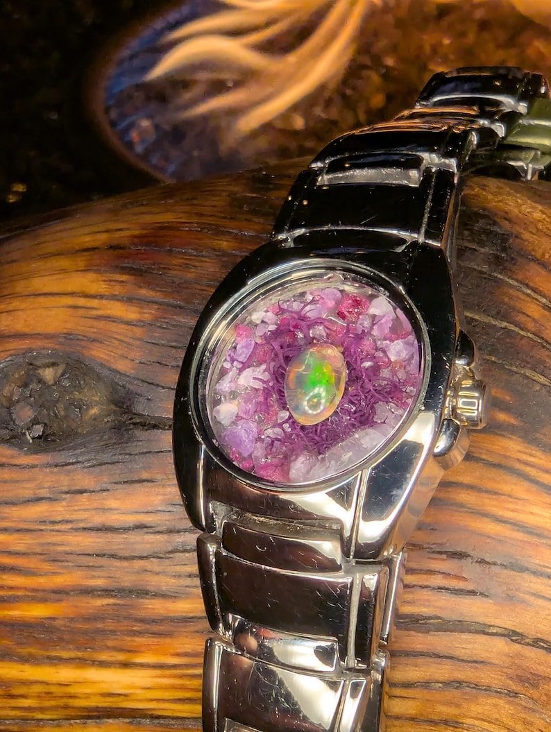 May include: A silver wristwatch with a unique face. The watch face is clear, revealing a colorful arrangement of small purple stones and a central opal. The opal has green and yellow hues. The watch band is also silver.