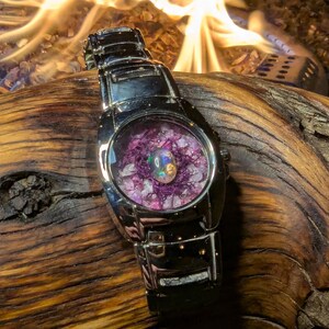 May include: A silver-toned wristwatch with a unique face. The watch face features a central opal surrounded by purple and white crystals. The watch is set against a backdrop of a wooden surface and a blurred fire.