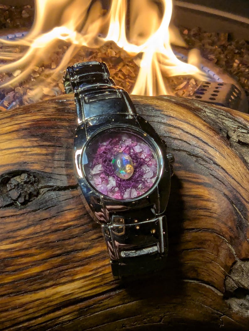 May include: A silver-toned wristwatch with a unique face. The watch face features a central opal surrounded by purple and white crystals. The watch is set against a backdrop of a wooden surface and a blurred fire.