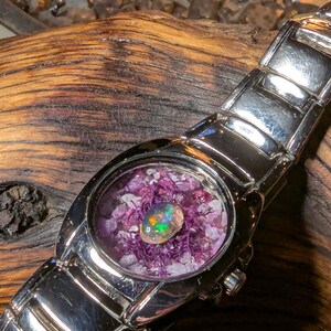 May include: A silver wristwatch with a unique face. The watch face features a colorful opal gemstone set in a bed of purple and white crystals. The watch band is made of polished silver links.