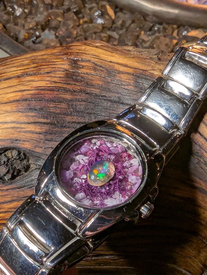 May include: A silver wristwatch with a unique face. The watch face features a colorful opal gemstone set in a bed of purple and white crystals. The watch band is made of polished silver links.