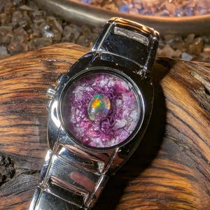 May include: A silver-toned wristwatch with a round face. The watch face features a colorful opal stone surrounded by purple and white crystals. The watch band is made of silver-toned metal.