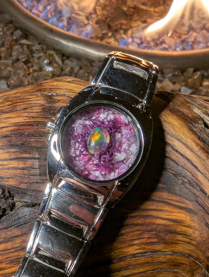 May include: A silver-toned wristwatch with a round face. The watch face features a colorful opal stone surrounded by purple and white crystals. The watch band is made of silver-toned metal.