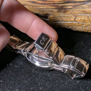 May include: A silver wristwatch with a rectangular link band. The watch face is round and the clasp is engraved with the letters "CR". The watch is displayed against a dark background.