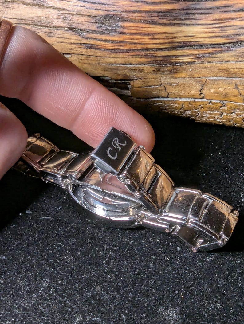May include: A silver wristwatch with a rectangular link band. The watch face is round and the clasp is engraved with the letters "CR". The watch is displayed against a dark background.