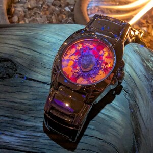 May include: A black wristwatch with a unique face design. The watch face features a colorful arrangement of red, orange, and purple elements. The watch is set against a dark, textured background.