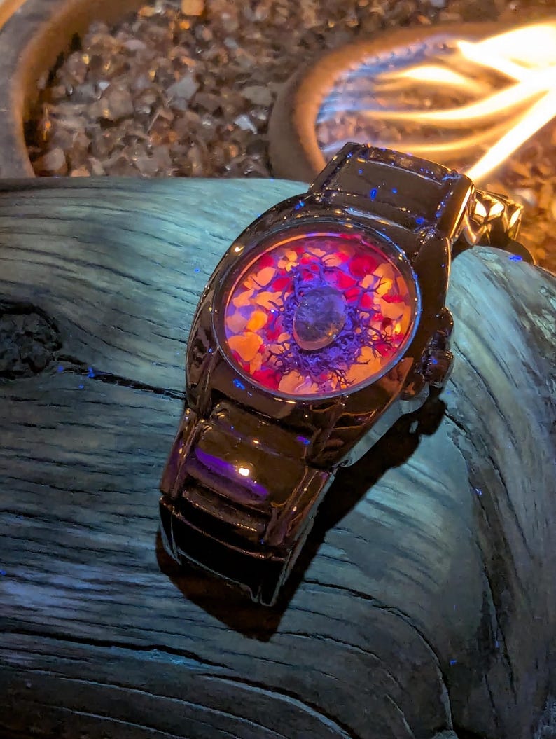 May include: A black wristwatch with a unique face design. The watch face features a colorful arrangement of red, orange, and purple elements. The watch is set against a dark, textured background.