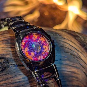 May include: A black wristwatch with a unique face design. The watch face features a colorful arrangement of orange, purple, and clear elements. The watch band is made of black metal. The background is blurred, with warm tones.