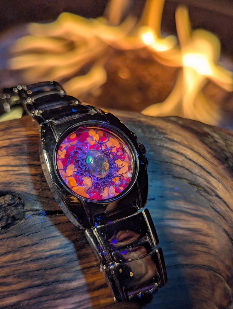 May include: A black wristwatch with a unique face design. The watch face features a colorful arrangement of orange, purple, and clear elements. The watch band is made of black metal. The background is blurred, with warm tones.