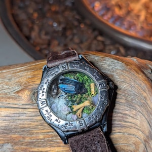 May include: A unique wristwatch with a black case and a brown leather strap. The watch face features a miniature diorama with a blue beetle, moss, and small bones. The bezel has compass markings. The watch is displayed on a piece of wood.