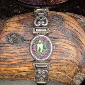 May include: A silver wristwatch with a unique design. The watch face features a white tooth set against a backdrop of green moss and purple flowers. The watch has a black leather band and ornate silver links.